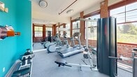 Located at 33 Railroad Avenue in Milford, Connecticut, this single-floor exercise room offers a well-equipped fitness space with a distinctive industrial character. The room is defined by its exposed red brick walls, which lend a warm, textured aesthetic throughout. Large, multi-pane windows with dark wood trim line the walls, flooding the space with abundant natural light and providing views of the surrounding greenery and streetscape — creating an energizing environment for workouts.

The flooring is covered in durable grey carpet, practical for a fitness setting. A vibrant teal accent wall on one side adds a bold pop of color, contrasting effectively with the brick and brightening the overall ambiance. Wall-mounted shelving and utility fixtures are present on this accent wall, alongside an electrical panel integrated neatly into the adjacent white wall.

The room is outfitted with a comprehensive range of fitness equipment, including treadmills, an elliptical machine, a multi-function cable/weight machine, a WaterRower rowing machine, a weight bench, and a dumbbell rack. Mirrors along one wall visually expand the space and assist users during strength training. Ceiling-mounted light fixtures provide ample illumination, and exposed red utility piping adds to the industrial aesthetic. The space is compact yet thoughtfully arranged to maximize functionality.