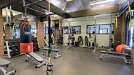 This single-level fitness space combines industrial aesthetics with a comprehensive range of training equipment. The interior is defined by its striking architectural character — exposed brick columns, sage-green painted walls, and a corrugated metal ceiling with integrated fluorescent lighting create a raw, functional atmosphere. Rubber flooring runs throughout, providing durability and safety across the training area.

The space is outfitted for a wide variety of workouts. Multiple squat racks with weight plates are positioned along the perimeter, alongside adjustable benches and a dumbbell rack catering to free weight training. TRX suspension trainers hang from the ceiling structure, offering bodyweight and functional training options, while battle ropes add to the conditioning equipment available. A heavy punching bag is visible near the dumbbell station, supporting combat fitness training.

Natural light enters through several windows positioned high along the walls, supplemented by the overhead fluorescent fixtures. The layout provides generous open floor space between equipment stations, allowing for movement and functional training. Full-length mirrors are integrated into the design, enhancing both the visual depth of the space and its practical use for form monitoring during workouts.