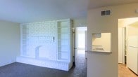 This single-floor space offers a thoughtfully laid-out interior that blends classic architectural character with functional design. The living area serves as the heart of the space, anchored by a striking white-painted brick fireplace that extends to the ceiling and is flanked by built-in open shelving — a standout feature that adds both charm and practical storage. The carpeted flooring throughout the living area lends a warm, comfortable feel underfoot. A doorway leads to an adjoining room that benefits from abundant natural light, hinting at a bright and airy atmosphere beyond. The kitchen is efficiently designed with a pass-through countertop that opens directly to the living space, creating a seamless, connected layout ideal for everyday living. The kitchen features light-toned cabinetry and countertops, as well as a sink positioned beneath an upper cabinet. A HVAC vent is integrated into the ceiling, ensuring year-round climate comfort. The textured ceiling adds a subtle layer of architectural detail throughout. Overall, the space presents a cohesive and practical layout with character-rich details that make it both livable and visually engaging.