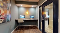 This single-story space features a dedicated computer workstation nook designed for focused productivity. The alcove is finished with cool gray-green walls that create a calm, professional atmosphere. A floating granite-topped counter spans the width of the nook, providing a sturdy and polished work surface that accommodates two desktop all-in-one computers, each accompanied by a sleek white barstool-height chair for comfortable seating.

Overhead, a distinctive yellow pendant light hangs from the ceiling, casting a warm ambient glow that complements the recessed lighting above. The combination of warm and cool lighting sources creates a well-lit environment suitable for extended computer use. The flooring features wood-look tile planks in a rich, dark tone, adding warmth and a contemporary aesthetic to the space.

Access to the workstation is provided through a full-length glass-panel door with a dark frame, allowing visibility into the adjacent area while maintaining a degree of separation. The overall design balances functionality with modern style, making it a practical and inviting workspace.