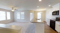 This single-floor home showcases an open-concept layout that seamlessly integrates the kitchen and living areas into one cohesive, light-filled space. The kitchen features a curved granite-topped island with an integrated sink, offering both a functional workspace and a natural dividing point between the kitchen and living zones. The flooring transitions from smooth hardwood-style surfaces in the kitchen area to plush carpeting in the living space, adding warmth and comfort underfoot.

Natural light fills the interior through multiple windows fitted with horizontal blinds, creating a bright and airy atmosphere throughout. The living area is equipped with a ceiling fan, while a chandelier above the dining area adds an elegant touch. Recessed lighting throughout the ceiling provides consistent, well-distributed illumination.

The neutral color palette — soft beige walls, white trim, and light-toned ceilings — contributes to a clean, versatile aesthetic that complements a variety of interior styles. A door with an adjacent window offers access to the exterior, further enhancing the home's connection to natural light. Overall, the space presents a practical and welcoming environment with modern finishes.