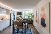 This thoughtfully designed single-floor unit showcases a contemporary open-concept layout that seamlessly connects the kitchen, dining, and living areas. The kitchen is outfitted with flat-front light blue cabinetry, under-cabinet LED lighting, a smooth cooktop, stainless steel appliances including a refrigerator and built-in microwave, and clean white countertops — offering both style and functionality. Rich wood-tone flooring runs throughout, grounding the space with warmth.

The dining area sits centrally between the kitchen and living room, defined by a patterned blue area rug and accompanied by a dark wood table with bar-height seating. The living area is anchored by a sectional sofa and benefits from expansive floor-to-ceiling windows that frame lush greenery and surrounding neighborhood views, filling the space with abundant natural light.

The bedrooms continue the modern aesthetic with crisp white walls, wood-tone flooring, and generous windows that bring in treetop views and outdoor scenery. Each bedroom offers a calm, well-lit retreat with ample space. Ambient cove lighting along the kitchen ceiling adds a refined architectural touch. Throughout, the unit maintains a clean, contemporary palette with pops of color from curated artwork and textiles, creating an inviting and cohesive atmosphere.
