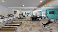 This single-level fitness center is designed to accommodate a wide range of workout styles, combining a spacious cardio and strength training area with a separate functional training zone. The space is unified by a distinctive geometric-patterned carpet in neutral tones accented with teal, lending a modern, energetic aesthetic throughout.

The main gym area is generously sized and outfitted with multiple treadmills, elliptical machines, and a comprehensive selection of strength equipment, including a full dumbbell rack and cable machines. Floor-to-ceiling mirrors line the walls, amplifying the sense of space and allowing users to monitor form. A wall-mounted flat-screen television adds convenience for cardio sessions. Recessed lighting throughout ensures the space is well-lit.

Adjacent to the main floor, a dedicated functional training room provides space for dynamic workouts. This area features wall-mounted suspension training equipment, a plyosoft box, stability balls, a hula hoop, and a step platform. Large windows in this zone invite natural light and offer views of the surrounding outdoor landscape. French-style glass doors provide access between areas, contributing to an open, connected feel across the entire fitness center.