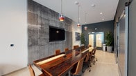 This single-floor space seamlessly combines industrial character with modern sophistication. The centerpiece of the conference area is a commanding live-edge wood table with resin inlays, surrounded by tan leather rolling chairs and illuminated by a series of distinctive red and copper cage pendant lights overhead. A raw exposed concrete feature wall serves as a dramatic backdrop, housing a wall-mounted flat-screen display ideal for presentations. Dark teal-painted walls contrast with light tile flooring throughout, creating a refined yet edgy atmosphere. Sliding barn-style doors on one wall offer flexible room division, while framed abstract artwork adds curated visual interest. A lounge area at one end provides a more relaxed setting, featuring mid-century-inspired armchairs, a low black credenza topped with an oversized industrial clock, and large windows that flood the space with natural light and offer views of an outdoor courtyard and pool area. Tall indoor plants are positioned throughout, softening the industrial elements and adding life to the space. Double glass-paneled entry doors flanked by lush potted greenery provide a welcoming transition point, completing this thoughtfully designed, multi-functional environment suited for meetings, collaboration, and informal gatherings.