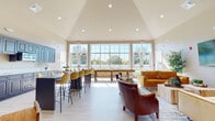 This single-floor community space is designed to accommodate a variety of social and recreational activities in a welcoming, modern environment. The centerpiece of the space is a generously sized open-concept lounge area anchored by soaring vaulted ceilings fitted with recessed lighting, creating a bright and airy atmosphere. A wall of large, grid-paned windows floods the interior with abundant natural light and offers views of the surrounding outdoor greenery.

The lounge is furnished with plush mustard-yellow sofas, a rich leather accent chair, and layered area rugs over light hardwood-style flooring, adding warmth and texture to the contemporary design. A large wall-mounted flat-screen television serves as a focal point in the seating area, complemented by decorative artwork and lush potted plants throughout.

A long bar-height counter with cushioned yellow barstools provides additional seating and a casual dining option, while a dedicated kitchenette features rich dark wood cabinetry, white countertops, and a microwave — ideal for hosting events. A separate flex area with round high-top tables and wood-and-metal stools offers a more intimate setting for small gatherings or casual meetings. A large barn-style sliding door adds an industrial-chic design detail to this versatile and well-appointed community space.
