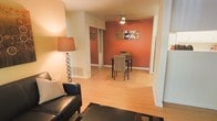 This single-floor space showcases an open-plan design that connects the living and dining areas in a cohesive, functional layout. The living area is anchored by a sleek black leather armchair, a patterned area rug, and a stylish floor lamp that casts warm ambient light, creating an inviting atmosphere. Hardwood-style flooring runs throughout the space, lending a clean, continuous aesthetic that ties the rooms together visually.

The dining area is defined by a bold terracotta-orange accent wall, which serves as a striking focal point and adds warmth and character to the interior. A compact dining table with cushioned chairs sits comfortably beneath a ceiling fan with integrated lighting, providing both ventilation and illumination. Decorative wall art above the dining table complements the rich wall color and enhances the overall design.

A sliding door visible in the background suggests access to an additional room or closet space, offering practical storage solutions. The neutral white walls in the living area balance the vibrancy of the accent wall, resulting in a space that feels warm yet airy. The overall ambiance is casual and comfortable, with thoughtful design details throughout.