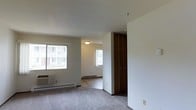 This single-floor space features a straightforward, functional layout with neutral tones and wall-to-wall carpeting throughout. The living area is bathed in natural light from multiple windows, offering views of the surrounding exterior. A wall-mounted air conditioning unit provides climate control, complemented by baseboard heating that runs along the perimeter of the rooms for year-round comfort. The open flow between the main living space and the adjoining room creates a sense of continuity, with a flush-mount ceiling light illuminating the secondary area. A full-height closet with classic folding panel doors offers built-in storage and serves as a defining architectural detail near the entryway. The clean white walls and low-profile ceilings contribute to a simple, utilitarian aesthetic throughout. The carpeted floors add warmth and comfort underfoot, while the neutral color palette provides a versatile backdrop. Overall, the space presents a practical and livable environment well-suited for everyday use.