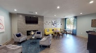 The IT Cafe is a thoughtfully designed single-floor communal space that seamlessly blends lounge comfort with café-style functionality. The interior is anchored by a striking stone-clad accent wall housing a sleek linear fireplace and a large wall-mounted flat-screen TV, creating a natural focal point for the main lounge area. Plush gray velvet armchairs arranged around a round coffee table on a teal area rug invite relaxed conversation and casual gatherings.

Adjacent to the lounge, a vibrant yellow banquette sofa with patterned accent pillows lines the wall beneath playful botanical wall art, paired with round wooden tables and wishbone-style chairs. A large arched window floods this dining nook with natural light and offers views of the surrounding greenery, framed by teal curtain panels that add a pop of color.

A separate bar-height workspace area features long dark wood communal tables and tall black stools beneath a warm pendant light, making it ideal for individual work or small group collaboration. This zone is accented with eclectic wall art and decorative pieces that contribute to the space's creative atmosphere.

Throughout, light wood-tone flooring, recessed lighting, and a neutral color palette with bold accent tones create a modern, welcoming ambiance suitable for both productivity and socializing.