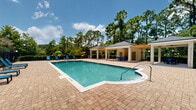 Located at 209 Cantabria Way in Saint Augustine, Florida, this outdoor community amenity area centers around a generously sized swimming pool with a maximum depth of 4 feet 6 inches, making it accessible for a wide range of swimmers. The pool itself features a classic rectangular shape with softened corners and is finished with clear, inviting turquoise water.

The surrounding deck is paved with attractive brick pavers, providing a durable and visually appealing surface throughout the entire pool area. Numerous blue lounge chairs are positioned along the pool's perimeter, offering ample space for sunbathing and relaxation.

Two covered cabana structures flank the poolside, each featuring ceiling fans, column supports, and dark shingled rooflines that provide shade and a comfortable outdoor gathering space. Seating and tables are arranged beneath each cabana, making them ideal for casual dining or socializing. A pergola element connecting the structures adds architectural interest and an additional shaded zone.

The entire area is enclosed by a black wrought-iron fence, ensuring privacy and security. A backdrop of tall pine trees and lush, mature landscaping creates a serene, natural atmosphere. The surrounding greenery provides both privacy and a park-like setting that enhances the overall outdoor experience of this well-appointed community amenity space.