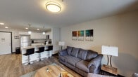 This single-floor unit is designed with a practical and modern open-concept layout, allowing the living, dining, and kitchen areas to flow seamlessly together. The living room is furnished with a plush gray sofa and armchair arranged around a round wooden coffee table, accented by a colorful triptych wall art piece and natural light from a large window. Wood-style flooring runs throughout the main living areas, adding warmth and continuity to the space.

The kitchen is well-appointed with white shaker-style cabinetry, granite-look countertops, and a full suite of stainless steel appliances, including a refrigerator, electric range, and microwave. A breakfast bar with stylish counter-height seating provides a casual dining option that connects directly to the living space.

The bedroom is generously sized and features carpet flooring, a wall-mounted television, a dresser, and direct access to a private balcony through sliding glass doors with vertical blinds. The bathroom includes a shower with a curtain enclosure, wood-style flooring, a white vanity, and direct access to a walk-in closet with built-in shelving, offering practical storage.