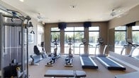 The fitness center is a thoughtfully designed, single-level space that combines functionality with an inviting atmosphere. Large floor-to-ceiling windows along the back wall flood the room with natural light and offer views of an outdoor pool area, creating an open, airy feel that enhances the workout environment. The warm, neutral-toned walls are accented by darker trim panels, giving the space a modern and polished aesthetic.

The room is equipped with a well-rounded selection of cardio machines, including treadmills, ellipticals, and stationary bikes — both upright and recumbent — arranged to allow comfortable spacing between users. Mounted flat-screen televisions are positioned above the windows, providing entertainment during workouts. A ceiling fan ensures air circulation throughout the space, contributing to a comfortable climate. Recessed lighting supplements the abundant natural light, keeping the room well-lit at any time of day.

A cable resistance machine is also available for strength training, broadening the facility's exercise options. The carpeted flooring adds a degree of comfort underfoot, while the overall layout ensures easy navigation between equipment, making this fitness center a practical and welcoming space for daily exercise.