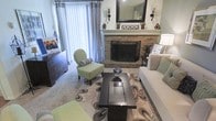 This charming single-floor space offers a thoughtfully designed interior that blends comfort and classic style. The living area serves as the heart of the home, centered around a striking brick fireplace crowned with a white mantel. A large rectangular mirror and decorative wall sconces above the mantel add a refined touch, while a traditional iron fireplace screen completes the look. Natural light filters into the room through full-length vertical blinds and elegant curtain panels, creating a bright and welcoming atmosphere. The space is furnished with a neutral-toned sofa accented with green and grey throw pillows, a charming sage green accent chair with curved wooden legs, and a sleek dark espresso coffee table resting atop a boldly patterned area rug. A dark wood dresser along one wall provides both storage and visual contrast to the lighter tones of the room. The soft carpeted flooring adds warmth underfoot and contributes to the overall cozy feel of the space. Wall-mounted sconces and table lamps provide layered lighting, enhancing the inviting ambiance. Every detail of this space reflects a careful balance of functionality and timeless interior design.