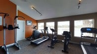 This single-floor fitness room is designed to support an active lifestyle with a focused and functional layout. The space is outfitted with a variety of cardio equipment, including a treadmill, elliptical trainer, upright stationary bike, and a recumbent bike, providing users with multiple options for cardiovascular exercise.

The room's interior design features a striking combination of a bold orange accent wall and warm taupe-toned walls, creating an energetic yet grounded atmosphere conducive to exercise. The textured white ceiling is fitted with track lighting, ensuring the space is well-illuminated at all times.

A row of large windows along one wall floods the room with generous natural light and offers views of the surrounding outdoor environment, enhancing the overall experience of the space. The flooring consists of dark, stone-patterned tile that is both durable and easy to maintain, well-suited for a high-traffic fitness environment.

A single door provides access to the room, keeping the layout clean and uncluttered. The combination of practical equipment, thoughtful design elements, and abundant natural light makes this fitness room a comfortable and motivating environment for daily workouts.