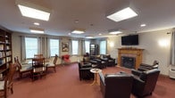 This single-floor communal space is thoughtfully designed to provide a warm and inviting environment for gathering and relaxation. The lounge area is anchored by a classic wood-mantled fireplace, which serves as a natural focal point and adds a sense of warmth and character to the room. A wall-mounted flat-screen television is positioned nearby, making the space well-suited for group viewing and entertainment.

Comfortable dark leather armchairs and sofas are arranged throughout the room to encourage conversation and relaxation, while additional seating options, including wooden and accent chairs, offer flexibility for various activities. A small side table provides a convenient surface for remotes and personal items.

The space is illuminated by a combination of large, flush-mounted ceiling light panels and soft wall sconces, ensuring even, welcoming lighting throughout. Multiple windows dressed with elegant floor-length curtains allow natural light to filter in, enhancing the open and airy feel of the room.

A snack and beverage vending machine is conveniently available along one wall, adding a practical amenity for occupants. Wall art and decorative accents contribute to the overall lived-in, homey ambiance. The flooring features wall-to-wall carpeting in a rich, deep tone, completing the comfortable and functional character of this communal retreat.