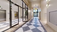 Located at 100 Place Charles-le Moyne in Longueuil, Quebec, the Ostral Lobby and Lounge is a thoughtfully designed communal space that makes a strong first impression. The entrance hallway is defined by a bold geometric checkerboard floor in grey and white, complemented by soaring ceilings and elegant pendant lighting, including a statement fringe chandelier that adds a warm, refined touch. Floor-to-ceiling black-framed glass doors and partitions flood the space with natural light while providing a sleek, contemporary aesthetic. Potted indoor trees add a refreshing organic element throughout.

At the heart of the lobby stands a curved wood reception desk beneath an arched wood-panel backdrop bearing the Ostral branding, anchoring the space with a welcoming focal point. A mailbox alcove and elevator access are conveniently integrated into the layout. The flooring transitions seamlessly from the patterned tile entryway to light wood-toned flooring as the space opens into the lounge area.

The lounge is a serene retreat furnished with a plush white sofa, round marble-topped coffee tables, and a built-in banquette seating nook accented by warm pendant lights and lush greenery. Open shelving along one wall adds a functional yet decorative element. The overall ambiance is calm, modern, and inviting — a polished communal space designed for comfort and style.
