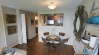 This single-floor unit offers an efficiently designed open-concept layout that seamlessly connects the main living areas. The dining area serves as a central focal point, accommodating a round dark-finished table paired with four matching chairs and a decorative candle centerpiece. Rich, dark hardwood flooring runs throughout the space, adding warmth and visual continuity.

The kitchen is positioned adjacent to the dining area and is accessible via a pass-through counter, which enhances the open feel while maintaining a defined cooking space. The kitchen features dark wood cabinetry, stainless steel appliances including a refrigerator and microwave, and track lighting mounted on the ceiling to illuminate the workspace effectively.

A hallway visible from the dining space leads to additional rooms, including what appears to be a bedroom with framed wall art, offering a sense of depth and connectivity throughout the unit. The walls are finished in a neutral gray-green tone, complementing the darker flooring and cabinetry. A textured ceiling and flush-mount light fixture complete the understated, functional aesthetic of this well-appointed space.