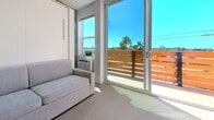 Located at 4245 Cleveland Ave in San Diego's vibrant Mission Hills neighborhood, this thoughtfully designed 160-square-foot studio maximizes every inch of its single-floor layout. The open-concept living area features a space-saving Murphy bed paired with a sofa, offering flexible use of the space. Large sliding glass doors flood the interior with abundant natural light and open directly onto a private balcony with warm wood-slat railings and open sky views of the surrounding San Diego cityscape, including palm trees and blue skies. A wall-mounted A/C unit ensures year-round comfort. The fully equipped kitchen runs along one wall and showcases butcher block countertops, white cabinetry with matte black hardware, a stainless steel range with overhead range hood, a double basin sink, and a white tile backsplash. A washer is also conveniently integrated into the unit. The entryway features exposed concrete block walls and a sleek black security door, adding an industrial-modern character to the space. A separate bathroom includes a glass-enclosed walk-in shower and toilet. Polished concrete flooring runs throughout, tying together the clean, contemporary aesthetic of this efficient and well-appointed urban studio.