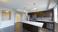 This single-floor space showcases a well-appointed kitchen designed with both style and functionality in mind. The kitchen is anchored by a large central island topped with light granite countertops featuring subtle veining, providing ample prep and gathering space. Dark espresso-toned cabinetry lines the walls, offering generous storage while creating a striking contrast against the light countertops and grey subway tile backsplash. Stainless steel appliances, including a gas range, built-in microwave, and French door refrigerator, contribute a polished, contemporary feel. Pendant lighting fixtures hang above the island and adjacent area, casting warm, focused light throughout the space. The hardwood flooring in a rich, dark tone runs throughout, adding warmth and cohesion. An open layout connects the kitchen to adjacent areas, enhancing the sense of flow. A glimpse through an open doorway reveals a warmly lit room beyond, suggesting comfortable living spaces nearby. The overall ambiance is modern and refined, balancing dark and light tones for a sophisticated yet inviting atmosphere.