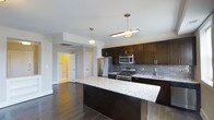 This single-floor space showcases a well-appointed kitchen designed with both style and functionality in mind. The kitchen is anchored by a large central island topped with light granite countertops featuring subtle veining, providing ample prep and gathering space. Dark espresso-toned cabinetry lines the walls, offering generous storage while creating a striking contrast against the light countertops and grey subway tile backsplash. Stainless steel appliances, including a gas range, built-in microwave, and French door refrigerator, contribute a polished, contemporary feel. Pendant lighting fixtures hang above the island and adjacent area, casting warm, focused light throughout the space. The hardwood flooring in a rich, dark tone runs throughout, adding warmth and cohesion. An open layout connects the kitchen to adjacent areas, enhancing the sense of flow. A glimpse through an open doorway reveals a warmly lit room beyond, suggesting comfortable living spaces nearby. The overall ambiance is modern and refined, balancing dark and light tones for a sophisticated yet inviting atmosphere.