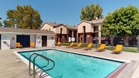 This outdoor amenity space centers around a generously sized swimming pool with clear, turquoise water, accessible via stainless steel entry rails and surrounded by a spacious concrete deck. The pool area is well-equipped for relaxation, featuring multiple bright yellow and orange lounge chairs arranged along the poolside, ideal for sunbathing under open skies. A dedicated pool house structure is positioned adjacent to the pool, providing utility and storage access for residents. The entire pool area is enclosed by a black metal fence, ensuring safety and privacy while maintaining an open, airy feel. Mature deciduous trees frame the perimeter of the space, offering natural shade and a lush backdrop that enhances the overall atmosphere. The surrounding multi-story residential buildings feature a warm earth-toned stucco and brick exterior with covered balconies and exterior staircases, creating a cohesive architectural character throughout the community. Well-maintained green lawns are visible beyond the fencing, contributing to the park-like setting. The space benefits from abundant natural light and clear, sunny skies, making it an inviting and functional outdoor retreat for residents throughout the warmer seasons.