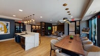 This single-floor amenity space is thoughtfully designed to serve multiple functions, blending social, recreational, and collaborative areas within one cohesive, open layout. Light hardwood flooring runs throughout, unifying the distinct zones while contributing to a warm, contemporary aesthetic.

The kitchen bar area anchors one end of the space, featuring dark navy cabinetry, a white quartz countertop, and a bold hexagonal tile backsplash. Amber leather barstools and decorative glass pendant lights add a stylish, inviting character. Adjacent to the bar, a long communal wood dining table with upholstered seating serves as a central gathering point, illuminated by cascading pendant fixtures overhead.

A lounge zone offers a sectional sofa, a wall-mounted flat-screen TV, and textured wallcovering, creating an intimate yet open retreat. A slatted dark wood partition with integrated shelving visually separates the games area, which features a billiard table beneath an industrial pendant light.

A dedicated conference room at the far end of the space is enclosed by floor-to-ceiling glass doors that open directly to an outdoor terrace, flooding the room with natural light and providing views of the exterior amenity area. The room accommodates a large rectangular meeting table with ample seating.