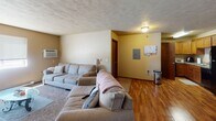 Located at 3200 S Theodore Ave in Sioux Falls, South Dakota, this single-floor residence offers a practical and comfortable layout. The living room is generously sized, featuring wall-to-wall carpet, a textured ceiling, and a large window that allows natural light to fill the space. A wall-mounted air conditioning unit provides climate control, and baseboard heating runs along the perimeter walls. The room offers ample space for a full sectional seating arrangement, making it well-suited for everyday living and entertaining. Adjacent to the living area is a carpeted hallway leading to interior doors, providing access to additional rooms. The kitchen is positioned near the front entryway and features warm-toned wood cabinetry with granite-look countertops, a black refrigerator, a dishwasher, and a sink with a window above it. The kitchen and entryway area are finished with hardwood-style flooring, creating a clean transition between spaces. A solid wood front door with a secure latch anchors the entryway. The overall layout is straightforward and functional, with neutral wall tones throughout that lend a calm, cohesive ambiance to the entire space.