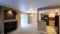 This single-floor space offers an open-concept layout that seamlessly connects the living room, dining area, and kitchen. The living room features neutral-toned carpet flooring and a built-in fireplace set within a dark-framed alcove, providing a cozy focal point. A sliding glass door floods the space with natural light and opens to an outdoor balcony with views of lush greenery beyond.

The dining area sits adjacent to the living room and is illuminated by a pendant chandelier, creating a defined yet connected space for entertaining. Warm beige walls complement the light-colored carpet throughout, maintaining a cohesive and welcoming ambiance.

The kitchen is partially open to the main living area via a raised bar counter with dark granite-style countertops, enhancing the sense of connectivity. It is equipped with dark appliances, including a black range and over-the-range microwave, and features medium-toned wood cabinetry. Recessed lighting ensures the kitchen remains well-lit. White baseboards and clean architectural details, including an arched passageway between spaces, contribute to the overall polished and functional character of the space.