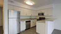 This single-floor space showcases a well-appointed kitchen designed with both functionality and aesthetics in mind. The kitchen features crisp white flat-panel cabinetry extending from floor to ceiling, providing generous storage capacity. Granite countertops with a subtle speckled pattern run along the perimeter, adding a natural, polished element to the space. Stainless steel appliances, including a refrigerator, dishwasher, and range, complement the clean, contemporary palette. A peninsula with a matching granite surface extends into the space, offering additional prep area and serving as a natural divider between the kitchen and the adjoining area. The undermount sink is paired with a modern pull-down faucet, positioned beneath upper cabinetry for practical workflow. Warm-toned hardwood flooring runs throughout, providing a grounding contrast to the white cabinetry and light walls. The overall design creates a cohesive, neutral aesthetic that balances practicality with a clean, modern sensibility suited to everyday living.