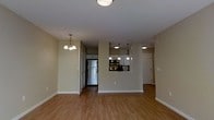 This single-floor residence offers a thoughtfully designed open-concept layout that seamlessly connects the living and kitchen areas. The space is finished with warm hardwood-style flooring that extends throughout, lending a cohesive and inviting feel to the interior.

The kitchen is a standout feature, outfitted with stainless steel appliances including a full-size refrigerator and built-in double wall ovens. White cabinetry provides ample storage, while pendant lighting suspended from the ceiling adds a modern touch above the kitchen pass-through. This open pass-through connects the kitchen to the main living area, creating an ideal layout for entertaining and everyday living.

The living area is generously proportioned, offering ample room for a variety of furniture arrangements. A classic chandelier with multiple candle-style lights serves as an elegant focal point overhead. The neutral, warm-toned walls throughout complement the flooring and contribute to the space's welcoming ambiance.

Additional doorways hint at further rooms beyond the main living space, including what appears to be a hallway leading to private quarters. White baseboards and clean wall finishes give the entire unit a polished, well-maintained appearance. Overall, this residence balances functional design with comfortable, timeless aesthetics.