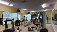 This fitness center offers a comprehensive workout environment designed to accommodate a range of exercise needs. The space is laid out on a single floor, making it easily accessible and straightforward to navigate. Mirrored walls line one side of the room, visually expanding the space while allowing users to monitor their form during workouts.

Natural light filters in through multiple windows fitted with blinds, complementing the overhead fluorescent lighting and ceiling fans that help maintain a comfortable atmosphere. The warm yellow-toned walls contribute to an inviting environment.

The cardio section features several machines, including ellipticals, a stationary bike, and a stair-climber or treadmill, each equipped with individual monitor screens mounted above for entertainment during workouts. A multi-station cable and weight machine anchors the strength training area, while a free weight rack with dumbbells and an exercise ball provide additional options. Rubber floor mats are placed in key workout zones for safety and comfort. Decorative indoor plants add a touch of greenery, softening the functional nature of the space and contributing to an overall welcoming, well-maintained atmosphere.