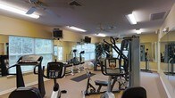This fitness center offers a comprehensive workout environment designed to accommodate a range of exercise needs. The space is laid out on a single floor, making it easily accessible and straightforward to navigate. Mirrored walls line one side of the room, visually expanding the space while allowing users to monitor their form during workouts.

Natural light filters in through multiple windows fitted with blinds, complementing the overhead fluorescent lighting and ceiling fans that help maintain a comfortable atmosphere. The warm yellow-toned walls contribute to an inviting environment.

The cardio section features several machines, including ellipticals, a stationary bike, and a stair-climber or treadmill, each equipped with individual monitor screens mounted above for entertainment during workouts. A multi-station cable and weight machine anchors the strength training area, while a free weight rack with dumbbells and an exercise ball provide additional options. Rubber floor mats are placed in key workout zones for safety and comfort. Decorative indoor plants add a touch of greenery, softening the functional nature of the space and contributing to an overall welcoming, well-maintained atmosphere.