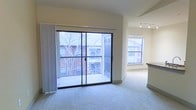 This single-floor residence offers a thoughtfully designed open layout that maximizes both functionality and comfort. The living area is carpeted and bathed in natural light, anchored by large sliding glass doors that open directly onto a private balcony with a metal railing — an ideal spot for taking in views of the surrounding neighborhood. Vertical blinds provide adjustable privacy and light control along the full width of the glass door. The transition from the living area into the kitchen is seamless, creating an open, connected feel throughout the common spaces. The kitchen area features hard flooring, a partial countertop or island visible at the edge of the frame, and a dedicated window that allows additional natural light to fill the space. Overhead track lighting in the kitchen provides focused, adjustable illumination. The neutral, warm-toned walls throughout maintain a clean and versatile aesthetic, suitable for a variety of interior styles. The overall ambiance of the residence is bright, airy, and welcoming, with an emphasis on natural light and open sightlines that lend the space a sense of openness and ease.