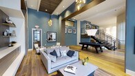 This two-story clubhouse is designed to impress, combining modern aesthetics with functional communal spaces that invite residents to gather and unwind. The main level features an expansive open-plan lounge area with soaring ceilings accented by rich wood beam detailing and a warm wood-toned ceiling inset. Bold teal-blue accent walls provide a vibrant backdrop to the contemporary furnishings, including a plush light-gray sofa, a sleek coffee table, and carefully curated artwork displayed along the walls. Eye-catching pendant lighting fixtures, including a cascading globe chandelier and a sculptural statement lamp, add an artful touch to the atmosphere. Wide-plank hardwood floors flow throughout, grounding the space with warmth and continuity. A full-size billiards table serves as a central entertainment feature, making the lounge ideal for social gatherings. A defined dining area is visible in the background, offering additional seating for residents. A modern staircase with cable railings leads to the second floor, which overlooks the main level, enhancing the sense of openness and connectivity between floors. The overall ambiance is upscale yet welcoming, blending residential comfort with boutique-style design.
