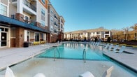 Located at 2301 E 10th St in Greenville, North Carolina, this outdoor amenity area serves as a central gathering space within a multi-story residential community. The centerpiece is a generously sized swimming pool with crystal-clear blue water, complemented by a shallow wading area featuring built-in water elements and sun loungers positioned directly in the water for relaxed sunbathing. The surrounding pool deck is finished in smooth concrete and offers ample space for lounging, furnished with bright yellow bench-style seating that adds a vibrant contrast to the neutral tones of the hardscape. A covered pergola structure provides shaded seating and serves as an elegant transition point between the pool deck and the surrounding landscape. The area is enclosed by wrought iron fencing, maintaining both security and an open, airy aesthetic. Brick and horizontal lap siding accent the exterior walls of the adjacent residential buildings, which rise several stories and feature private balconies overlooking the pool courtyard. Glass-paneled entry doors with brick surrounds provide convenient access to the pool area from the building interior. Landscaped greenery and manicured shrubs frame the perimeter, softening the hardscape and enhancing the overall ambiance. The space enjoys abundant natural light under open skies, making it an inviting outdoor retreat for residents.