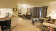 This single-floor space offers a thoughtfully arranged open-concept layout that seamlessly connects the living and dining areas, creating a sense of generous, flowing space. The living area is anchored by plush, neutral-toned carpeting that extends throughout, providing warmth and comfort underfoot. A distinctive glass-top coffee table with a sculptural dark wood base serves as a centerpiece, adding a touch of modern elegance to the lounge space.

The dining area is defined by a classic wooden dining table surrounded by matching dark wood chairs, ideal for hosting gatherings. Above, an elegant chandelier casts a warm, inviting glow over the space. The dining area is framed by bold, patterned floor-to-ceiling curtains in earth tones, which add texture and visual depth to the room while softening the overall aesthetic.

A partial wall with a countertop passthrough connects to an adjacent kitchen area, enhancing the open and communicative flow between spaces. Decorative touches, including potted plants and framed wall art, bring life and personality to the interiors. The warm, neutral color palette throughout creates a cohesive and inviting ambiance, making this space both functional and welcoming for everyday living and entertaining.