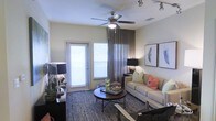 This single-floor space offers a thoughtfully designed interior with a comfortable and contemporary aesthetic. The living area serves as the heart of the space, showcasing a neutral color palette accented with vibrant pops of color through decorative pillows in coral, green, and cream tones. A plush sofa anchors the seating arrangement, complemented by a round wooden coffee table resting atop a bold chevron-patterned area rug that adds visual texture to the room. Overhead, a ceiling fan with a brushed nickel finish provides both function and style, while a track lighting system offers additional illumination and flexibility. Large windows dressed with white horizontal blinds and floor-length gray curtains flood the space with natural light, creating a bright and airy atmosphere. Framed botanical and nature-inspired artwork adorns the walls, contributing to the refined yet approachable ambiance. A dedicated accent corner features stylish table lamps and decorative objects, adding depth and personality to the overall design. The space strikes a balance between modern sophistication and everyday livability, making it both visually appealing and highly functional.