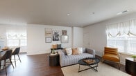 This single-floor residence is designed with a practical open-concept layout that connects the main living spaces seamlessly. The living room is generously proportioned, featuring rich dark hardwood flooring, neutral white walls, and a well-lit environment enhanced by natural light from windows with sheer draping. A curated gallery wall and an entry bench area add character to the space.

The kitchen is a functional highlight, outfitted with warm-toned wood cabinetry, granite countertops, stainless steel appliances, and a central island with bar seating for two. A dedicated pantry provides additional storage, and a window above the counter brings in natural light.

The bedrooms are carpeted for comfort, each offering ample space. The primary bedroom includes a window with outdoor views and room for a sitting area, while a secondary bedroom connects directly to a well-appointed bathroom. The bathroom features dark wood vanity cabinetry with granite countertops, a walk-in glass-enclosed shower with white tile surround, and hardwood-style flooring.

Overall, the residence presents a cohesive, contemporary aesthetic with warm wood tones, neutral finishes, and a thoughtfully organized floor plan suited for comfortable everyday living.