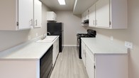 Located in Las Vegas, Nevada, this single-story space offers a thoughtfully designed interior highlighted by a galley-style kitchen. The kitchen is laid out with parallel runs of cabinetry on both sides, maximizing storage and counter space within an efficient, corridor-style layout. White shaker-style cabinets with brushed silver hardware line both walls from floor to ceiling, providing ample storage. Light-toned laminate countertops run continuously along both sides, offering generous workspace. Black appliances, including a refrigerator, electric range, and dishwasher, provide a clean contrast against the white cabinetry. A single-basin sink with a chrome faucet is integrated into one countertop run. An under-cabinet range hood is positioned above the stove for ventilation. The flooring throughout the kitchen consists of light wood-look planks, contributing to a clean, contemporary aesthetic. Overhead, recessed fluorescent lighting ensures the space is well-lit. The neutral gray walls complement the overall palette, creating a cohesive and practical interior environment suited to everyday living.