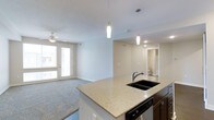 This single-floor residence showcases an open-concept layout that seamlessly connects the kitchen and living area, creating a spacious and functional environment. The kitchen is appointed with granite countertops, dark cabinetry, a black double-basin sink, and stainless steel appliances. A peninsula extends into the living space, providing additional prep space and a natural dividing point between the two areas. Overhead, two sleek pendant lights hang above the counter, adding a modern design touch.

The living area features neutral-toned carpet, white walls, and a ceiling fan, lending the room a clean, comfortable feel. Glass-paneled doors open to an outdoor space, allowing natural light to fill the room and offering exterior views. Recessed lighting is present throughout the space, supplementing both natural and pendant light sources.

A hallway visible from the main living area leads to what appears to be a bathroom and additional rooms, suggesting a well-organized floor plan. The overall aesthetic is contemporary and neutral, making the space adaptable to a variety of interior styles.