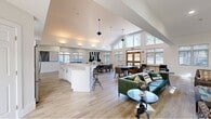 This generously sized single-floor community clubhouse offers an open-concept layout designed to accommodate a variety of activities and social gatherings. The most striking architectural feature is the soaring vaulted ceiling at one end of the main hall, flanked by a dramatic wall of floor-to-ceiling windows and a set of double glass doors that open to an outdoor patio, flooding the interior with natural light and views of the surrounding greenery.

Light wood-tone flooring runs throughout, complementing the crisp white walls and lending a clean, modern aesthetic. The main gathering area features multiple dining and lounge seating arrangements, including plush green sofas, upholstered chairs, and dark dining tables with cushioned chairs — ideal for both casual socializing and structured meetings.

A standout kitchen island with a marble-style quartz countertop serves as a central hub, complete with bar seating and backed by a well-appointed kitchen featuring white shaker cabinetry, marble tile backsplash, stainless steel appliances, and ample counter space.

A dedicated workstation area offers dual computer desks, comfortable seating, and a foosball table for recreation, all bathed in natural light from multiple windows. A cozy lounge corner with a marble-surround fireplace and wall-mounted television provides a warm, inviting retreat within the space.