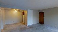 This single-floor unit offers an open and functional layout designed for efficient everyday living. The main living area is finished with wall-to-wall carpeting in a neutral tone, creating a soft and understated foundation throughout the space. A solid wood entry door provides a straightforward point of access, while a built-in closet with white bifold doors offers convenient storage directly within the living area.

The layout opens visually into the kitchen, which is partially visible from the main room. The kitchen features warm wood-toned upper cabinetry and includes a white refrigerator, suggesting a compact but practical cooking space. A ceiling fan with a light fixture is mounted overhead in the kitchen area, providing both ventilation and ambient lighting.

The walls are finished in a soft gray tone, giving the space a clean and neutral appearance throughout. The open sightlines between the living area and kitchen contribute to an airy feel despite the modest scale of the unit. Overall, the space presents a straightforward, no-frills interior that prioritizes functionality and ease of use.