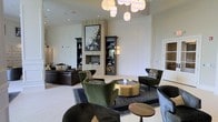This single-floor space exudes refined sophistication through its thoughtfully curated interiors and striking architectural elements. The dining area commands attention with its dramatic, branching globe chandelier suspended from soaring ceilings, casting a warm glow over a substantial dark stone dining table surrounded by sleek, transparent ghost chairs. Structural columns frame the space elegantly, while classic wainscoting and decorative wall panels add a tailored, timeless quality. A tufted banquette seating area anchors the far wall, complemented by wall-mounted accent lighting and curated artwork.

The adjacent lounge area is equally impressive, centered around a refined fireplace flanked by dark built-in shelving units displaying carefully chosen décor pieces. A large-scale abstract artwork serves as a focal point above the mantle, surrounded by a travertine surround and herringbone tile inset. Plush green velvet armchairs and a cylindrical brass coffee table create an inviting conversational seating arrangement, while an organic cluster chandelier overhead adds a sculptural element. Rich area rugs, layered textures, and a cohesive neutral palette with deep accent tones unify both spaces into a seamlessly flowing, upscale environment ideal for entertaining.