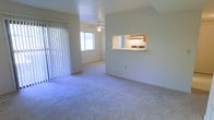 This single-floor residence offers a welcoming and functional layout designed for comfortable everyday living. The living area is spacious and open, finished with soft, plush wall-to-wall carpeting that adds warmth underfoot. A ceiling fan with a built-in light fixture anchors the room, providing both circulation and ambient lighting. Natural light flows generously through a large window dressed with vertical blinds, which also appears to lead to an outdoor area via a sliding glass door — bringing a bright, airy quality to the interior. A distinctive pass-through window connects the living space directly to the kitchen, creating an open sightline that enhances the sense of connectivity between the two areas. The kitchen, visible through the pass-through, features cabinetry and countertop appliances, offering a practical and efficient cooking space. The neutral wall tones throughout contribute to a clean, versatile ambiance that is easy to personalize. The overall design balances openness with a cozy residential feel, making the space feel welcoming and move-in ready.