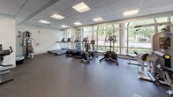 The fitness center at 138 College is a single-level facility designed to support a comprehensive workout experience. The space is finished with durable rubber flooring throughout, providing both safety and functionality across all exercise zones. A dropped ceiling with recessed panel lighting ensures consistent, even illumination throughout the entire area.

One of the facility's most notable features is a large floor-to-ceiling window wall that lines a significant portion of the space, flooding the interior with natural light and offering open views of the surrounding greenery outside. This bright, airy atmosphere creates an inviting environment for exercise.

The cardio section is well-appointed with multiple treadmills, elliptical trainers, and a stationary bike, all positioned to take advantage of the outdoor views. The strength training area includes a multi-function cable machine, a leg extension and curl machine, and additional selectorized equipment. A three-tier dumbbell rack holds a full range of free weights, complemented by medicine balls and a wall-mounted mirror for form feedback. A wall-mounted flat-screen television is positioned above the free weight area. Glass double doors provide entry to the facility, with a water station and waste bin conveniently located near the entrance.
