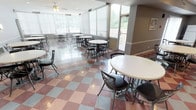 This single-floor communal space is designed to accommodate group gatherings and dining, offering a welcoming and functional environment throughout. The main dining area is generously proportioned, featuring a distinctive checkerboard-patterned tile floor in shades of red, gray, and beige that adds a classic character to the space. Multiple round tables with metal chairs are arranged throughout, providing flexible seating for a variety of group sizes.

Large windows line the perimeter of the dining area, flooding the interior with abundant natural light and offering views of the surrounding greenery. Vertical blinds and roll shades provide adjustable light control. A ceiling fan with a light fixture is centrally positioned overhead, complementing the recessed lighting and wall-mounted sconces that illuminate the space.

An adjacent sunroom-style area extends the dining space, featuring expansive floor-to-ceiling windows and direct access to an outdoor area, creating a seamless connection between the interior and exterior. This area carries the same tile flooring and round table seating, making it ideal for casual gatherings.

The space also includes a functional kitchen equipped with a range, oven, microwave, and ample counter space along a light blue cabinetry island. The kitchen connects directly to the dining area, supporting catered events or communal meal preparation. Overall, this space is practical, bright, and well-suited for community use.