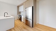 This modern 2-bedroom, 2-bathroom unit at 2800 Boulevard Thimens in Montréal offers a thoughtfully designed open-concept layout with clean lines and contemporary finishes throughout. Light-toned hardwood flooring flows seamlessly across the living spaces, enhancing the bright, airy atmosphere created by large windows that invite abundant natural light and outdoor views of surrounding greenery.

The kitchen is a standout feature, showcasing a large central island with a white quartz countertop, a black matte sink fixture, white upper cabinetry, and warm wood-toned lower and tall cabinets that extend to the ceiling. Stainless steel appliances, including a built-in oven and full-size refrigerator, complete the functional and stylish space. The open living area is generously proportioned, with a pendant light fixture and dedicated in-unit stacked washer and dryer.

The two bedrooms are well-sized and finished with the same warm hardwood flooring and crisp white walls, with windows providing views of the exterior. Both bathrooms are elegantly appointed: one features a walk-in glass shower with large-format stone-look tiles and matte black fixtures, while the second includes a soaker tub/shower combo with matching tile surround. A sleek white vanity with black hardware adds to the refined, minimalist aesthetic throughout.