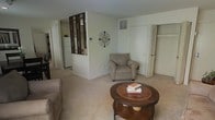 This single-floor residence offers a warm and functional layout designed for comfortable everyday living. The living room serves as the heart of the home, featuring wall-to-wall carpeting that adds a soft, inviting feel underfoot. A distinctive decorative lattice-style room divider separates the living area from the kitchen beyond, providing a charming architectural detail that maintains an open sense of flow while defining each space. The kitchen, visible through the divider, is equipped with standard appliances including a refrigerator. The living room is finished with neutral-toned walls that create a bright and airy ambiance, complemented by a ceiling vent that supports climate control throughout the space. A closet with bifold doors provides convenient storage directly off the living area. Wall decor, including a decorative metal accent piece, adds character to the otherwise clean and understated interior. The layout is straightforward and efficient, making the most of the available space on a single level. Overall, the home presents a tidy, move-in ready environment with a calm, neutral palette suited to a variety of furnishing styles and personal tastes.