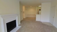 This single-floor space offers an open-concept layout that seamlessly connects the living area to the kitchen. The living room is carpeted in neutral beige tones, creating a warm and comfortable atmosphere. A white-mantled fireplace with a black surround serves as a focal point along one wall, adding a classic architectural detail to the space. The room is generously proportioned, providing ample space for furniture arrangement and everyday living.

The open layout transitions into a dedicated dining or flex area, delineated by a partial wall and column detail that adds architectural interest without closing off the space. A white breakfast bar separates this area from the kitchen, which is visible in the background. The kitchen features light wood cabinetry and includes a full-size refrigerator, illuminated by overhead fluorescent lighting that brightens the work area.

The walls throughout are painted in a soft, neutral cream tone, contributing to an airy and cohesive aesthetic. A utility panel is recessed into one wall, maintaining a clean, uncluttered look. Overall, the space presents a functional and inviting layout suited for comfortable everyday living.