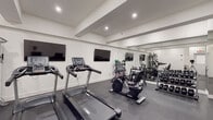 This fitness center is designed with a clean, modern aesthetic that supports a focused and functional workout environment. The space features crisp white walls, a coffered ceiling with recessed lighting, and durable dark rubber flooring that provides both traction and cushioning throughout.

The cardio zone is anchored by multiple Spirit-brand treadmills and stationary bikes, including a recumbent cycle and an upright model, all positioned in front of two large wall-mounted flat-screen televisions for entertainment during exercise. Adjacent to the cardio equipment, a Peloton-style spin bike rounds out the cycling options.

The strength training area is comprehensively outfitted with a full rack of hex dumbbells spanning a wide range of weights, a multi-station cable and resistance machine, an adjustable workout bench, and a selection of medicine balls and stability balls stored on a dedicated rack. Stacked exercise mats are also available for floor-based workouts and stretching.

Full-length mirrors line one wall, visually expanding the space and allowing users to monitor their form during exercise. The layout is thoughtfully arranged to provide ample open floor space between equipment zones, ensuring comfortable movement throughout. A clearly marked exit and fire safety equipment are also present, reflecting a commitment to safety within the facility.