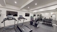 This fitness center is designed with a clean, modern aesthetic that supports a focused and functional workout environment. The space features crisp white walls, a coffered ceiling with recessed lighting, and durable dark rubber flooring that provides both traction and cushioning throughout.

The cardio zone is anchored by multiple Spirit-brand treadmills and stationary bikes, including a recumbent cycle and an upright model, all positioned in front of two large wall-mounted flat-screen televisions for entertainment during exercise. Adjacent to the cardio equipment, a Peloton-style spin bike rounds out the cycling options.

The strength training area is comprehensively outfitted with a full rack of hex dumbbells spanning a wide range of weights, a multi-station cable and resistance machine, an adjustable workout bench, and a selection of medicine balls and stability balls stored on a dedicated rack. Stacked exercise mats are also available for floor-based workouts and stretching.

Full-length mirrors line one wall, visually expanding the space and allowing users to monitor their form during exercise. The layout is thoughtfully arranged to provide ample open floor space between equipment zones, ensuring comfortable movement throughout. A clearly marked exit and fire safety equipment are also present, reflecting a commitment to safety within the facility.